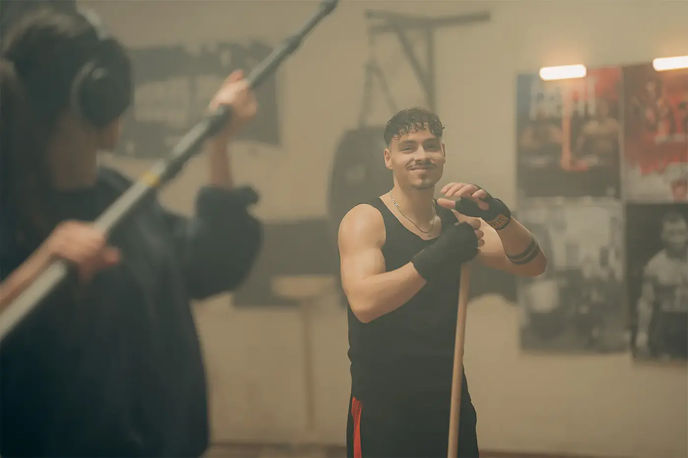 Lächelnder junger Mann lehnt an einem Besen im Boxstudio, Tonangel im Vordergrund, Standbild aus dem IAF Abschlussfilm Opernboxer 2025
