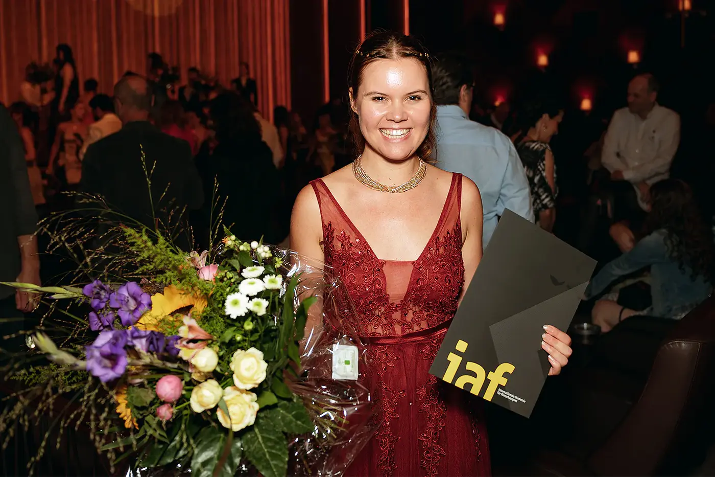 Absolventin im roten Spitzenkleid mit Blumenstrauß und IAF Zeugnismappe, IAF Abschlussfeier 2024 in Köln