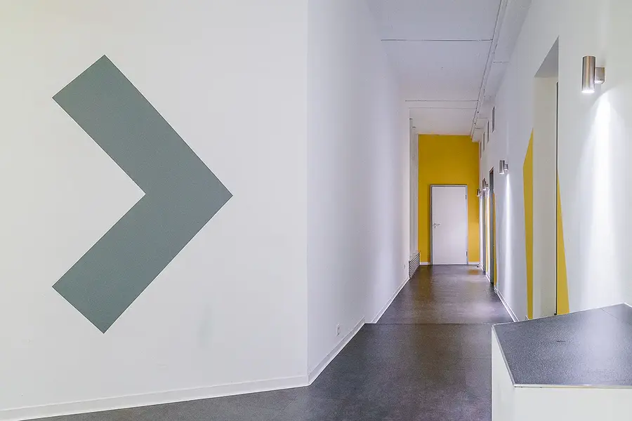 Flur der Filmschauspielschule mit grauem Pfeilsymbol und gelber Akzentwand